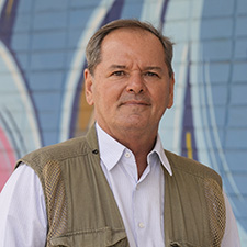 Sérgio Almeida, presidente da Lente Cultural.Homem branco, cabelo curto de cor castanho claro, de camiseta branca, e colete bege; posando em uma parede grafitada.