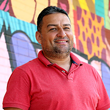 Marcelo Pereira. Edição de Vídeo. Homem branco, cabelo curto castanhos escuro, barba, de camiseta vermelha, posando em uma parede grafitada.