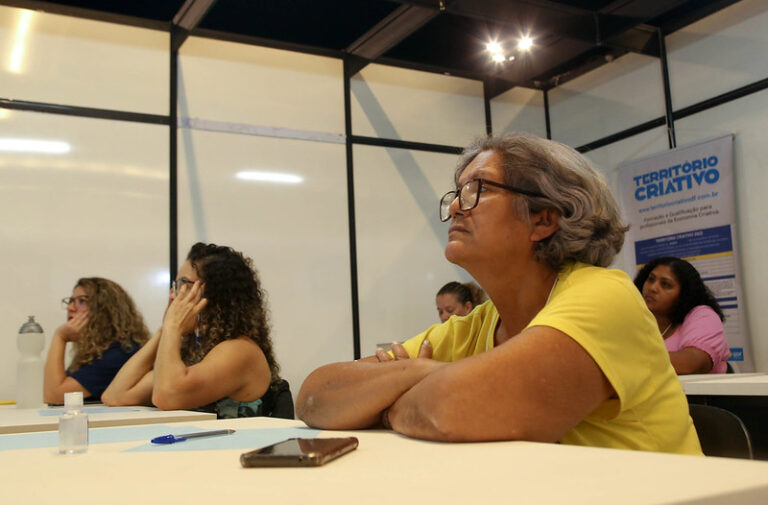 quatro mulheres sentadas em mesas de sala de aula. Em primeiro plano, uma mulher vestida de amarelo e usando óculos, cabelos grisalhos.